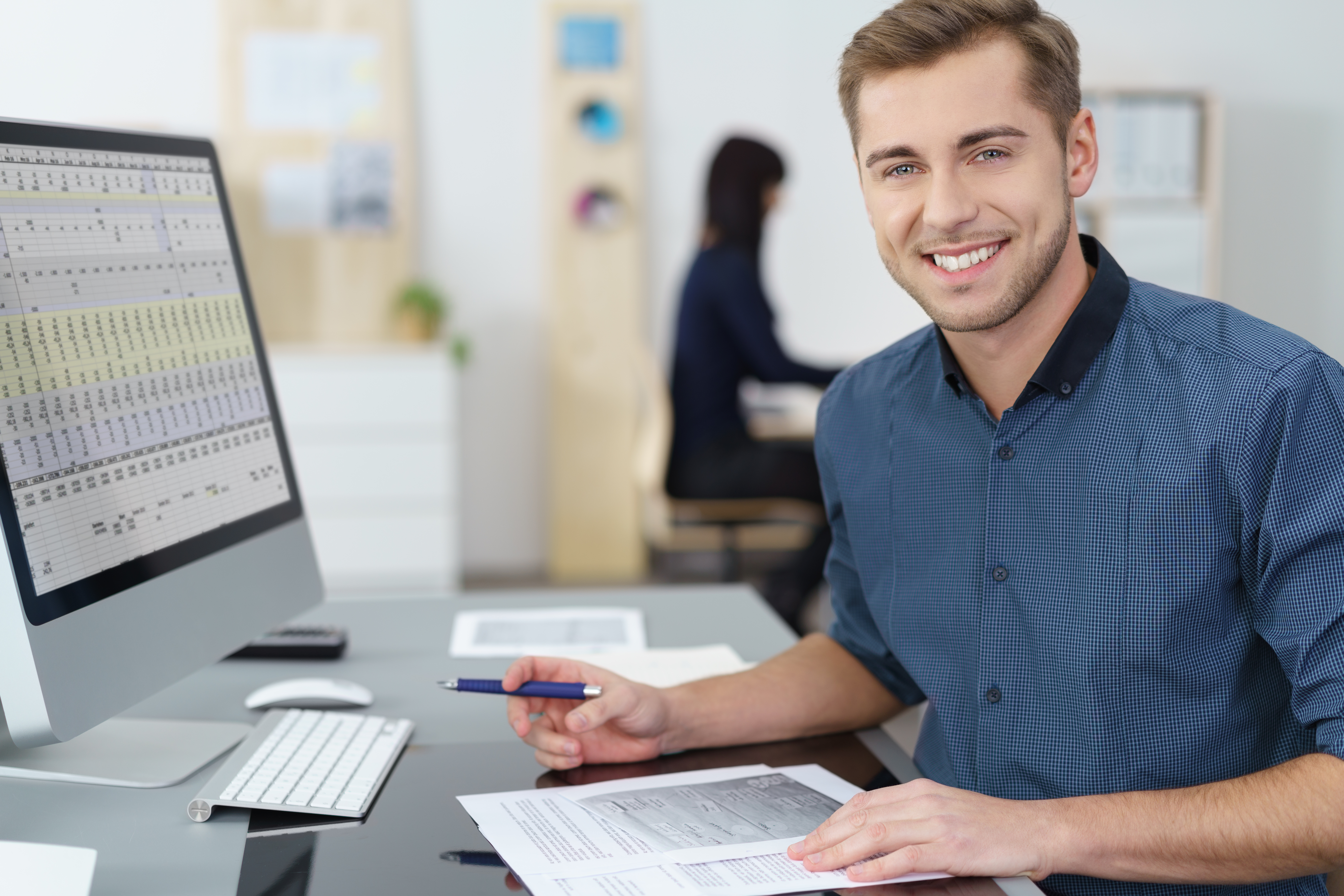 Ein Mann sitzt lächelnd an einem Schreibtisch mit Monitor und hält einen Stift in der Hand